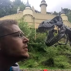 Visitor standing near a dinosaur model with a castle-style building in the background.