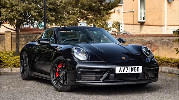 Black Porsche 911 convertible with red brake calipers, parked on tarmac in front of houses and trees, viewed from the front.