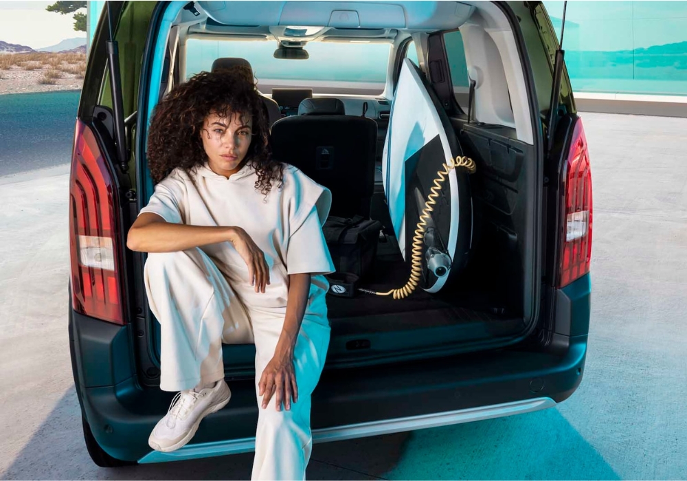 Woman sitting in the open cargo area of a Peugeot E-Rifter with a surfboard stored inside, in an outdoor setting.