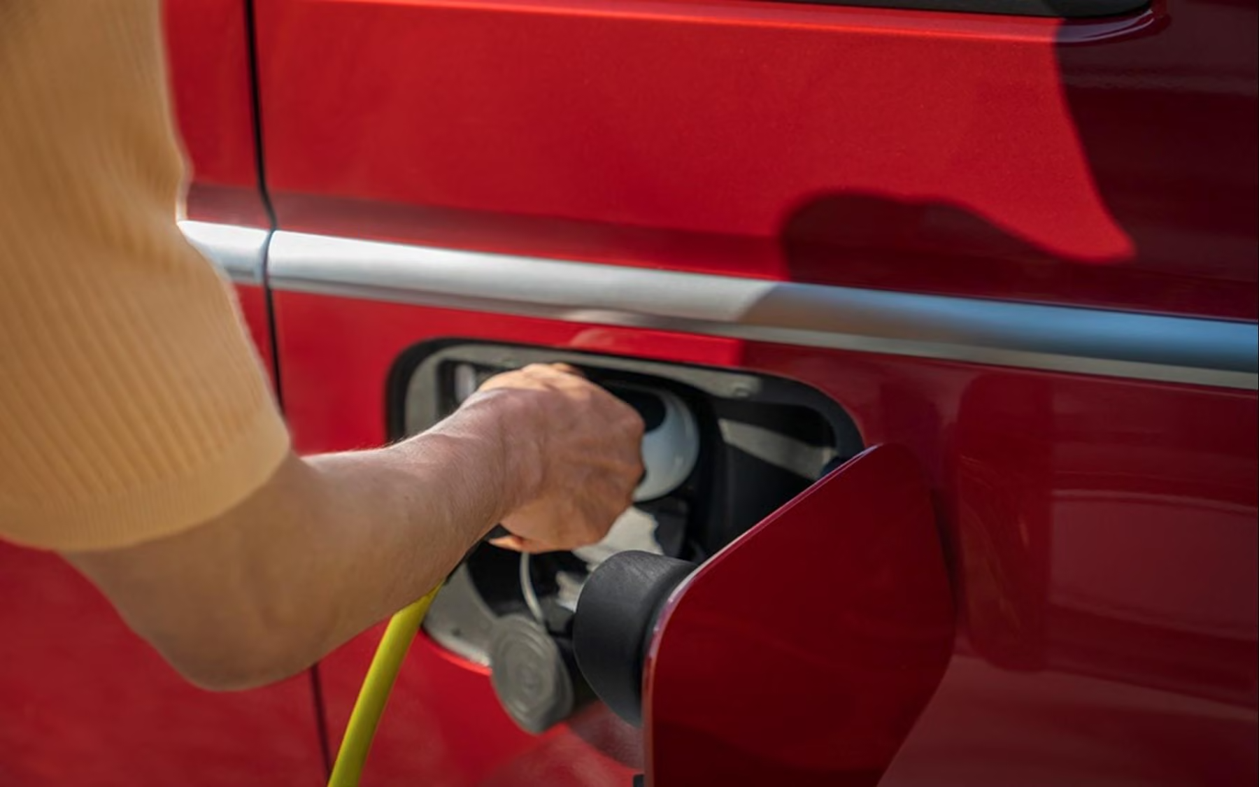 Close-up of a hand plugging a yellow charging cable into the charging port on the side of a red Volkswagen Multivan eHybrid, with the charge flap open