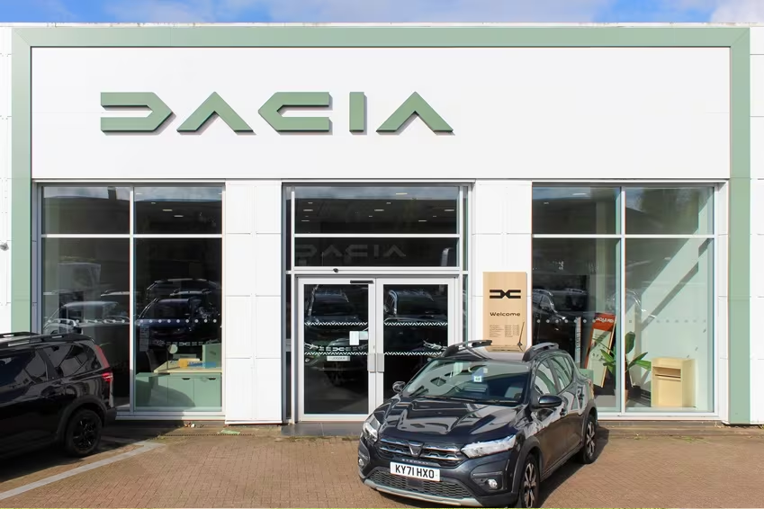 Front entrance of Dacia dealership with glass façade and parked Dacia vehicle in foreground.