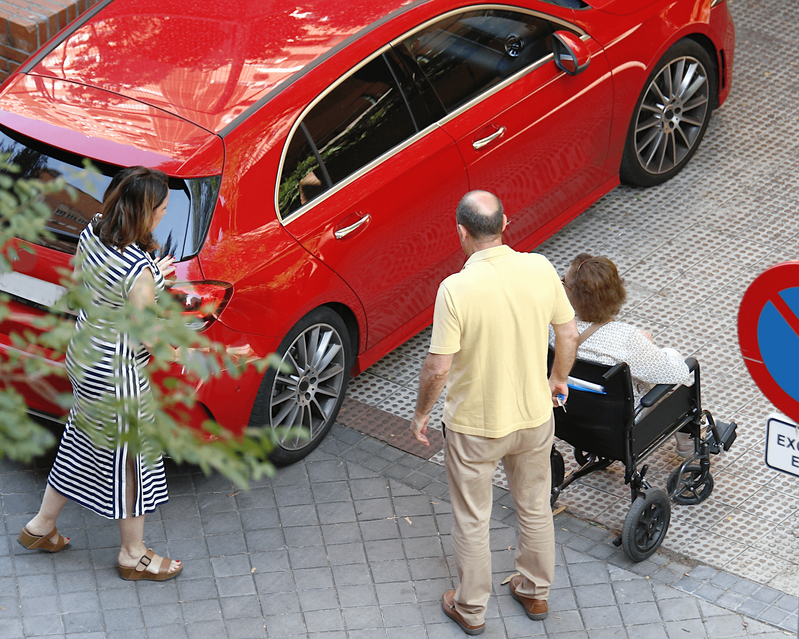 Person in wheelchair being assisted towards a red car parked on a street with two companions nearby.