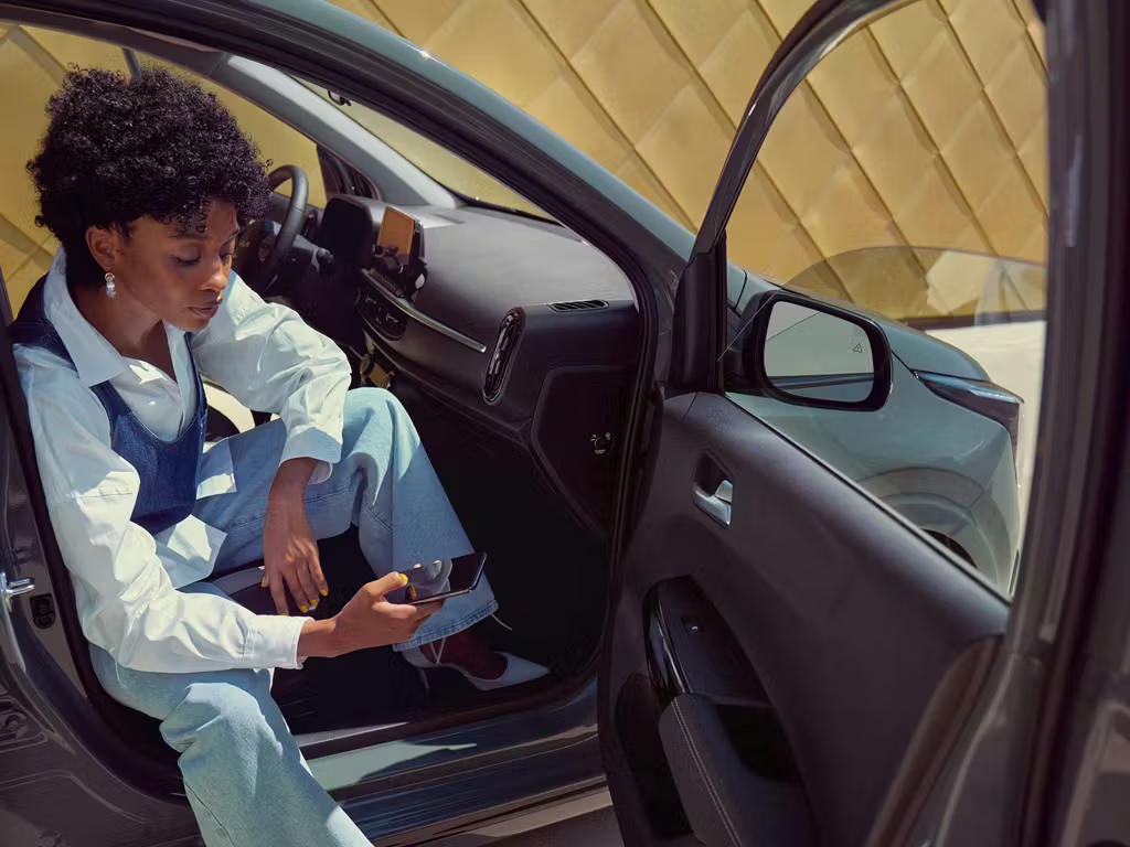 Woman seated in the driver's seat of a Kia Picanto with door open, using smartphone, showcasing the compact interior with dark trim and modern dashboard layout.