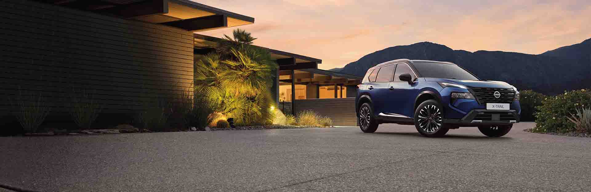 A blue nissan x trail driving on a snowy mountain path next to a lake.