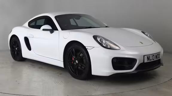 White Porsche Cayman coupe with black wheels, parked in a studio against a plain grey background, shown from the front three‑quarter view.