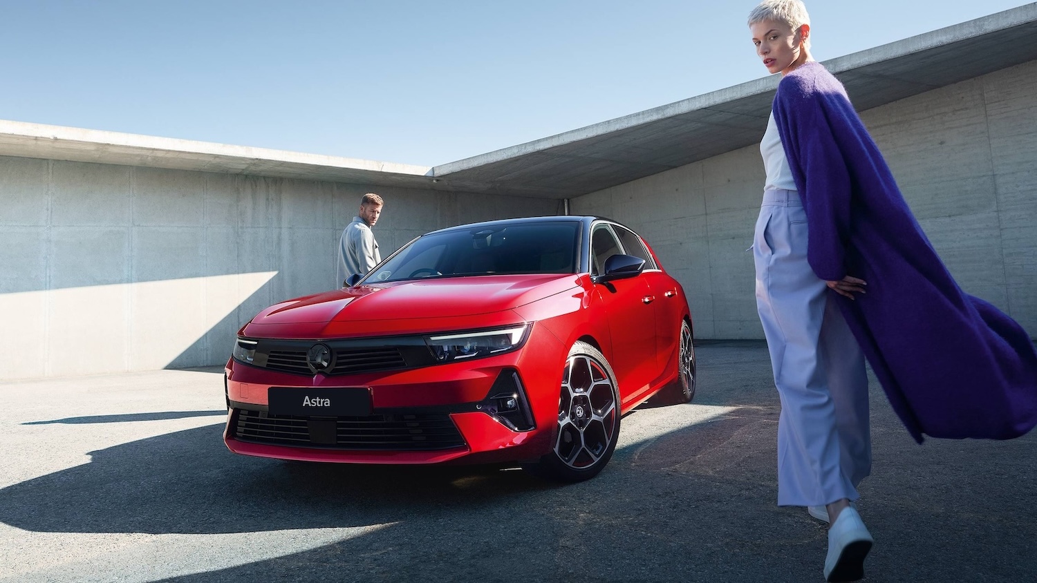Red Vauxhall Astra parked in a modern concrete setting with two people walking nearby.