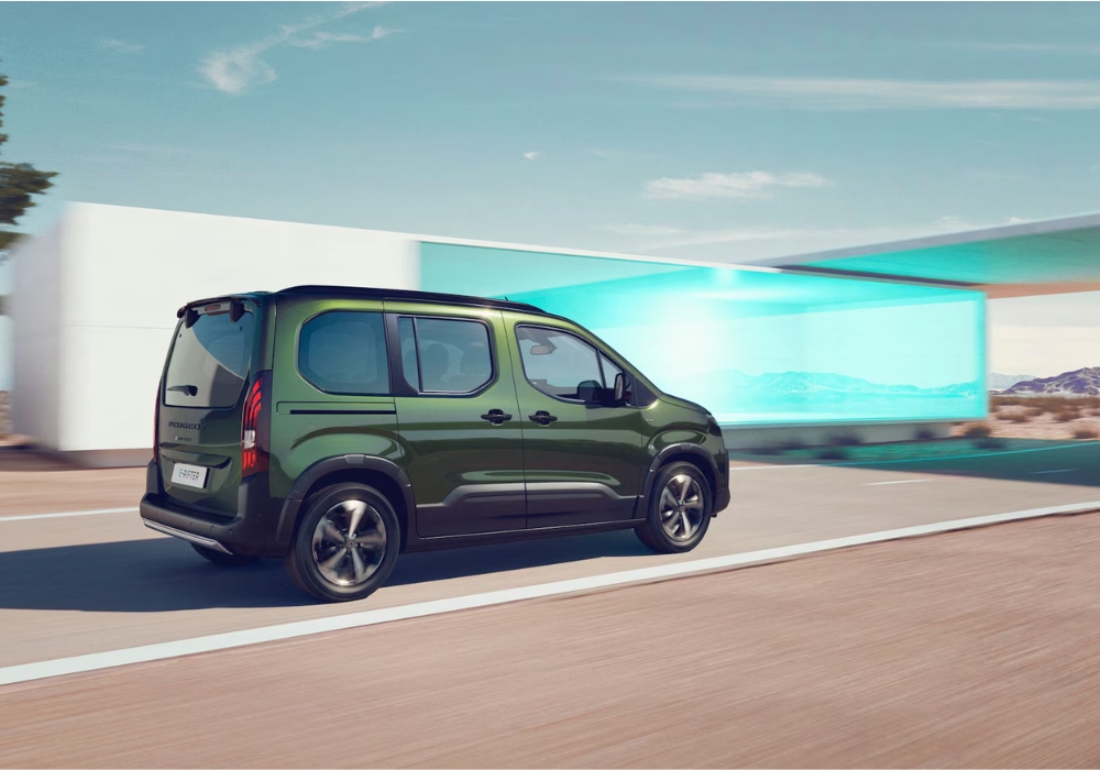 Peugeot E-Rifter in green shown from rear three-quarter angle driving on a road past a modern building.