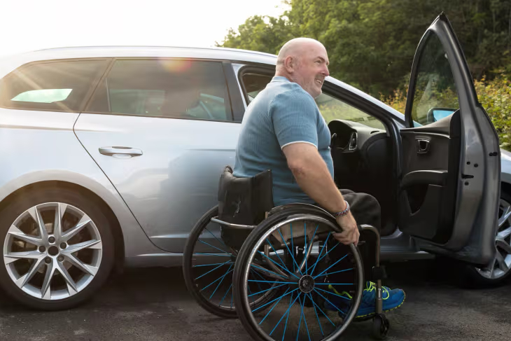 Person using a wheelchair transferring from a silver estate car with the driver’s door open, demonstrating accessible vehicle use.