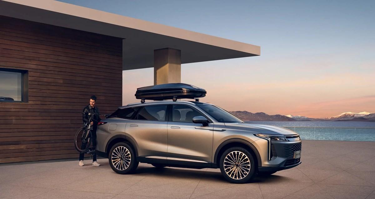 Omoda SUV with roof box parked outside a modern house by the coast, with a person standing beside a bicycle.