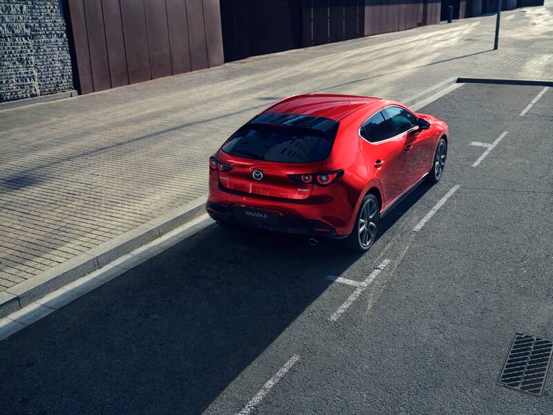 The rear of a red Mazda3 hatchback, parked in a parking space on the side of a road.