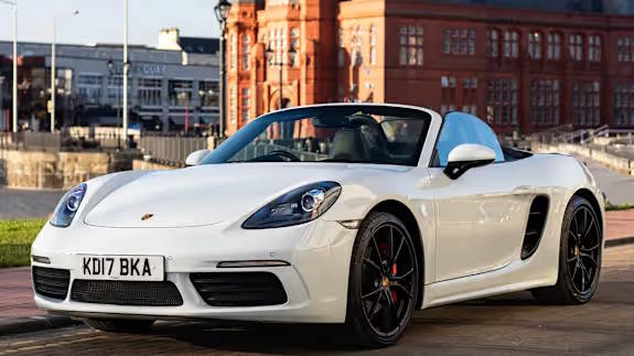 White Porsche 718 Boxster convertible with black wheels, roof down, parked on a quayside with red brick buildings and water in the background, viewed from the front three‑quarter angle.
