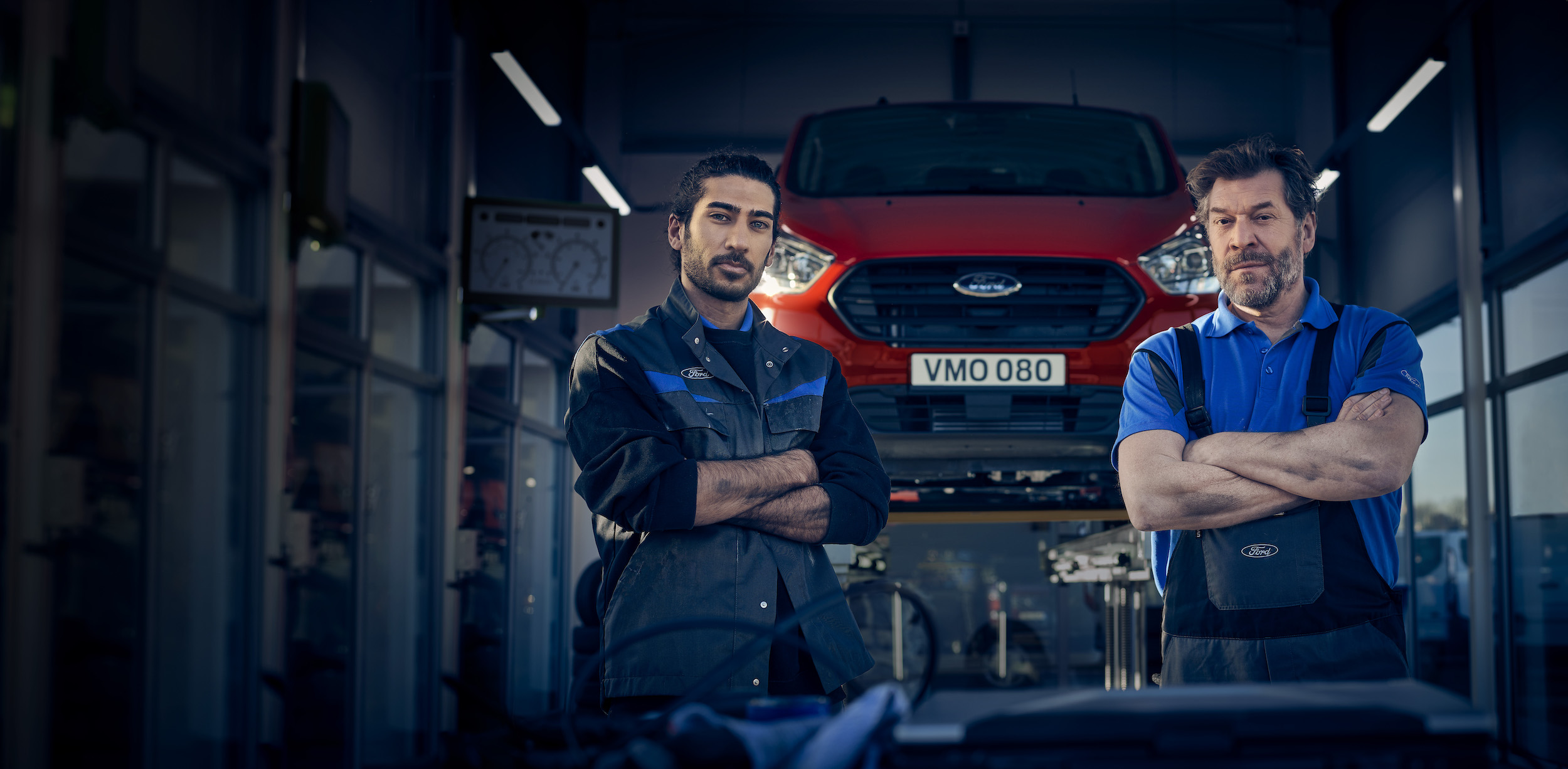 Two automotive technicians standing in a workshop with a red Ford vehicle on a lift behind them, arms crossed, looking at the camera.
