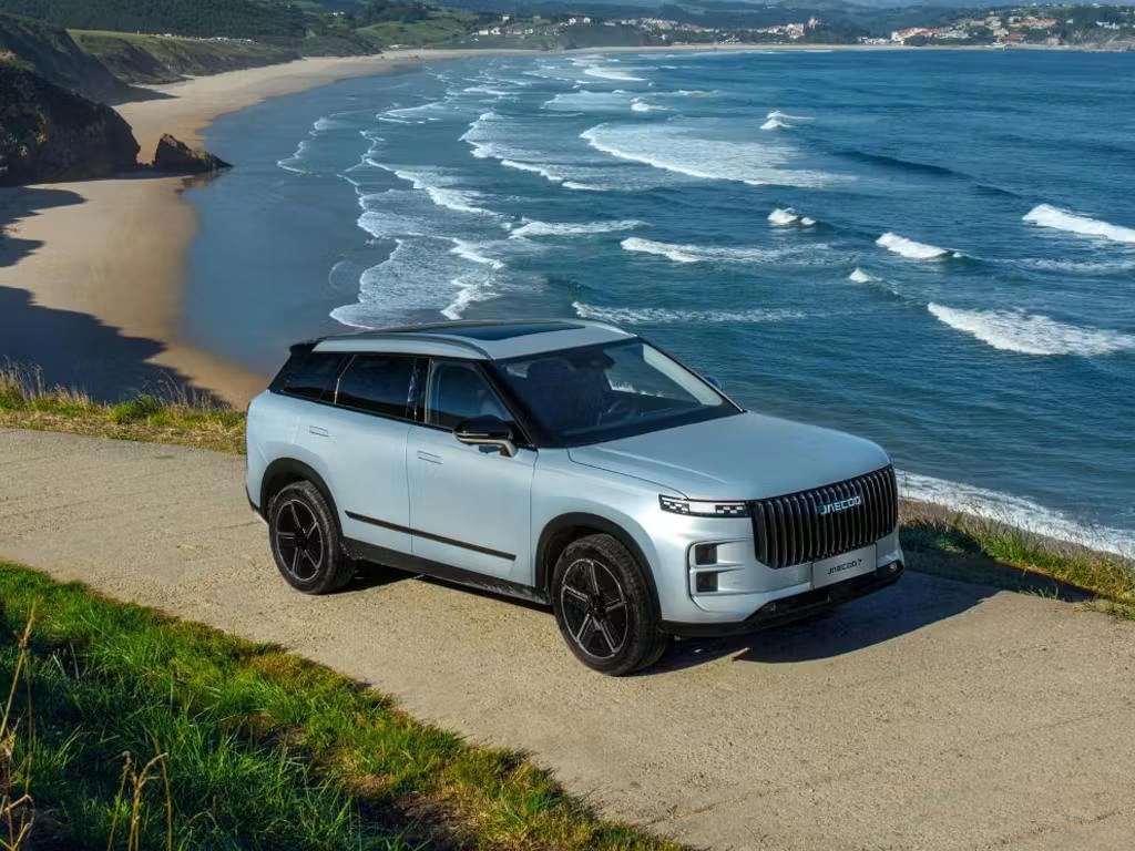 Front three-quarter elevated view of the Jaecoo 7 SUV in silver, featuring its bold vertical grille, sleek LED headlights and dark alloy wheels against a coastal backdrop.