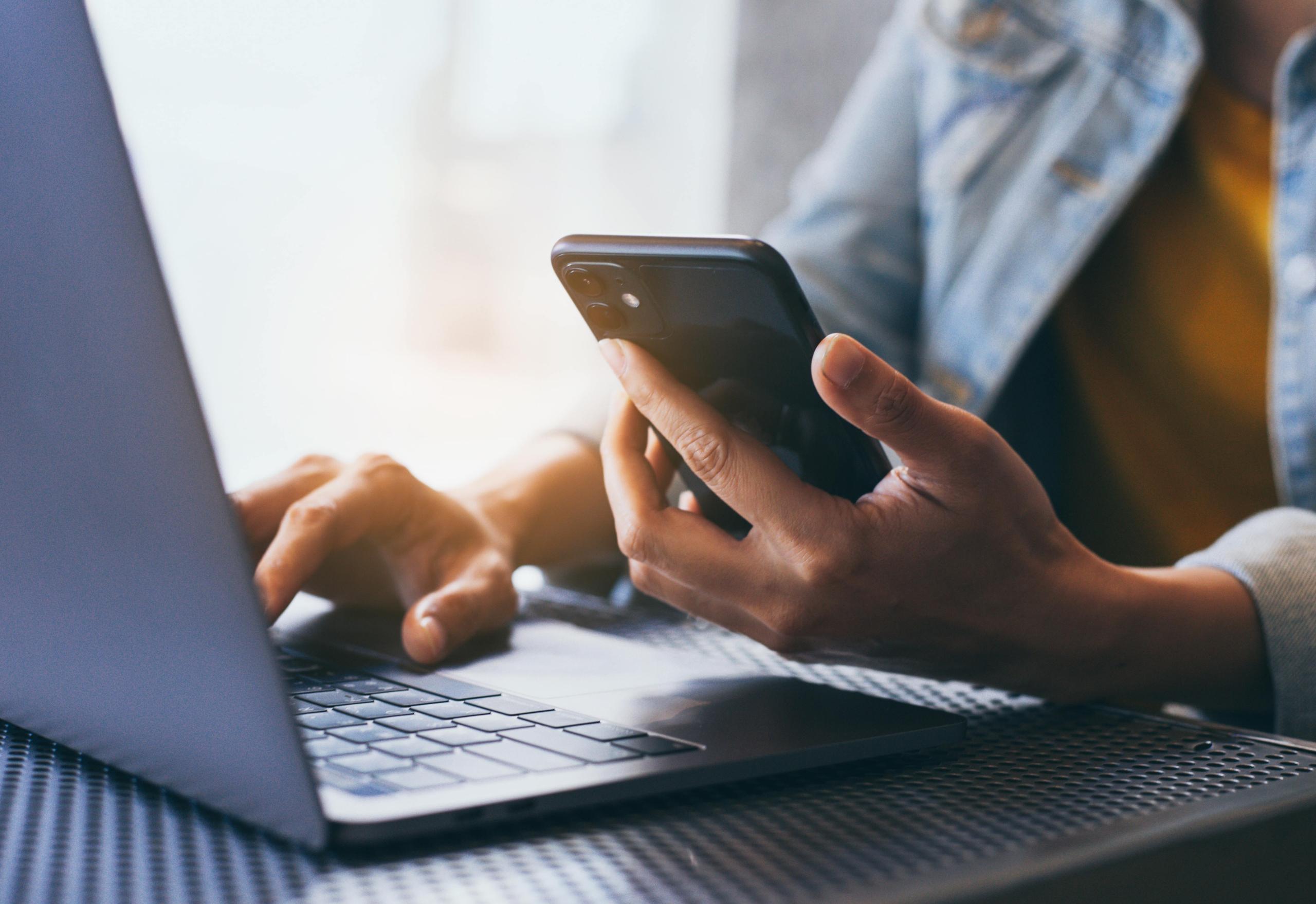 Woman using iPhone with one hand and typing on a macbook with the other.