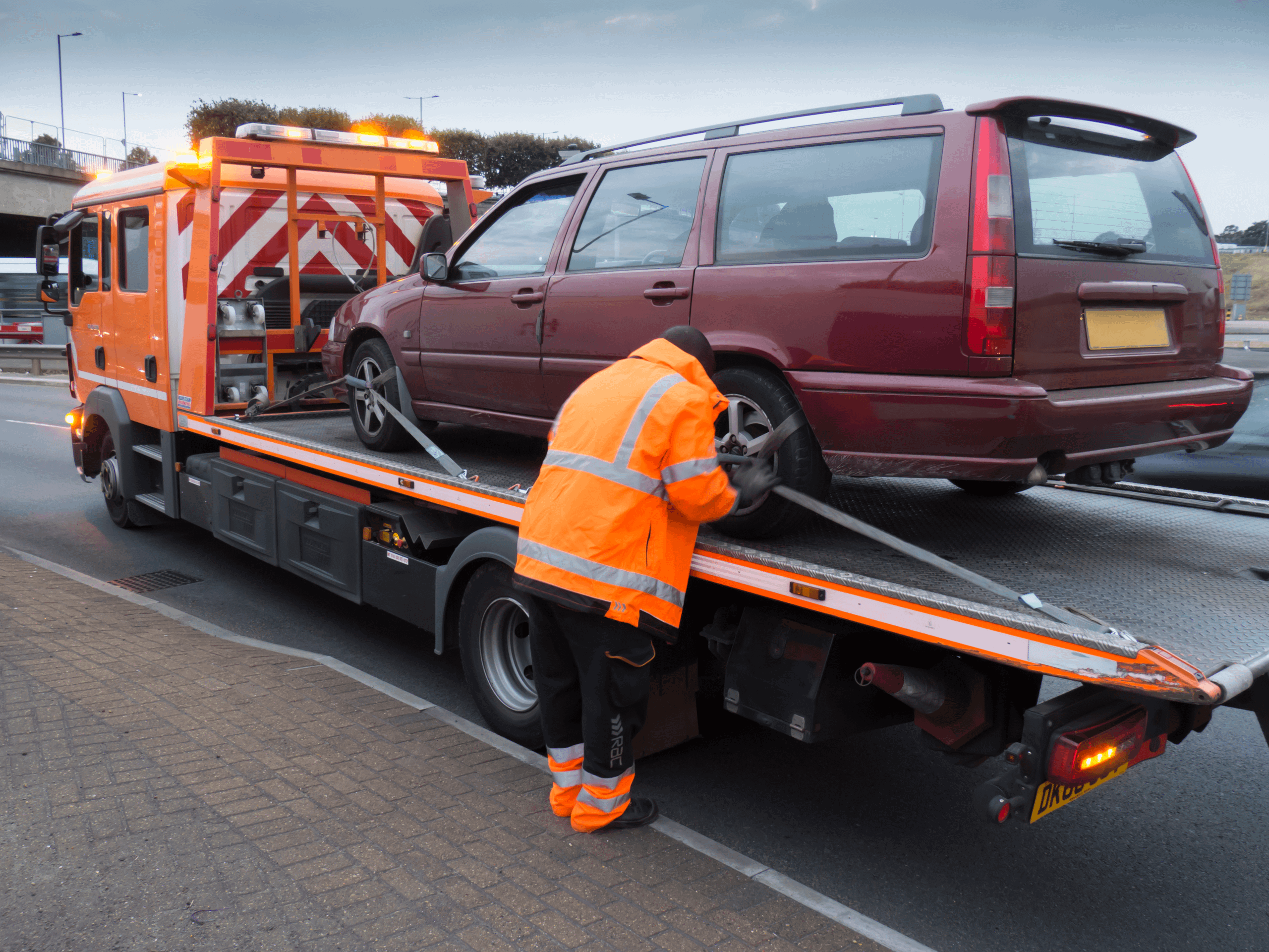 RAC worker in Orange Hi Vis wear securing a broken down red car onto a recovery wagon.