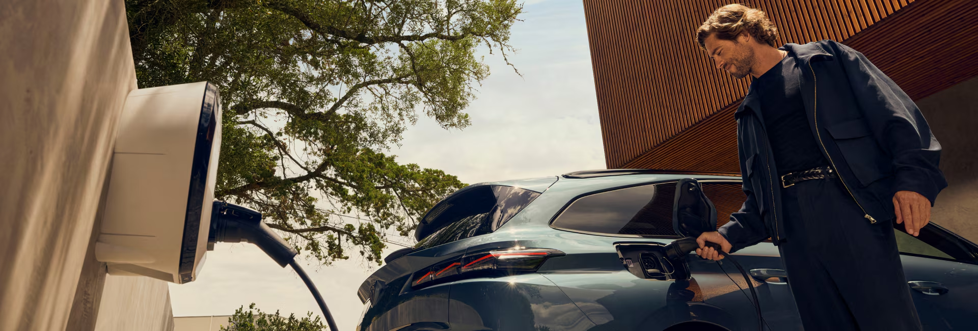 Man plugging in an electric vehicle to a wall-mounted home charging station beside a modern house, with trees and natural light in the background.