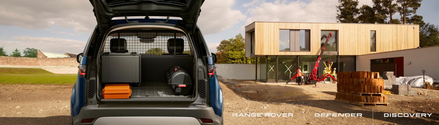 Rear cargo area of a Land Rover with the tailgate open showing tools and equipment, parked outside a modern house under construction.