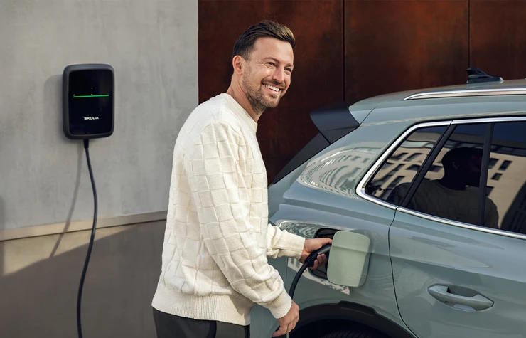 Smiling man charging a green Skoda SUV at a wall‑mounted home EV charger.