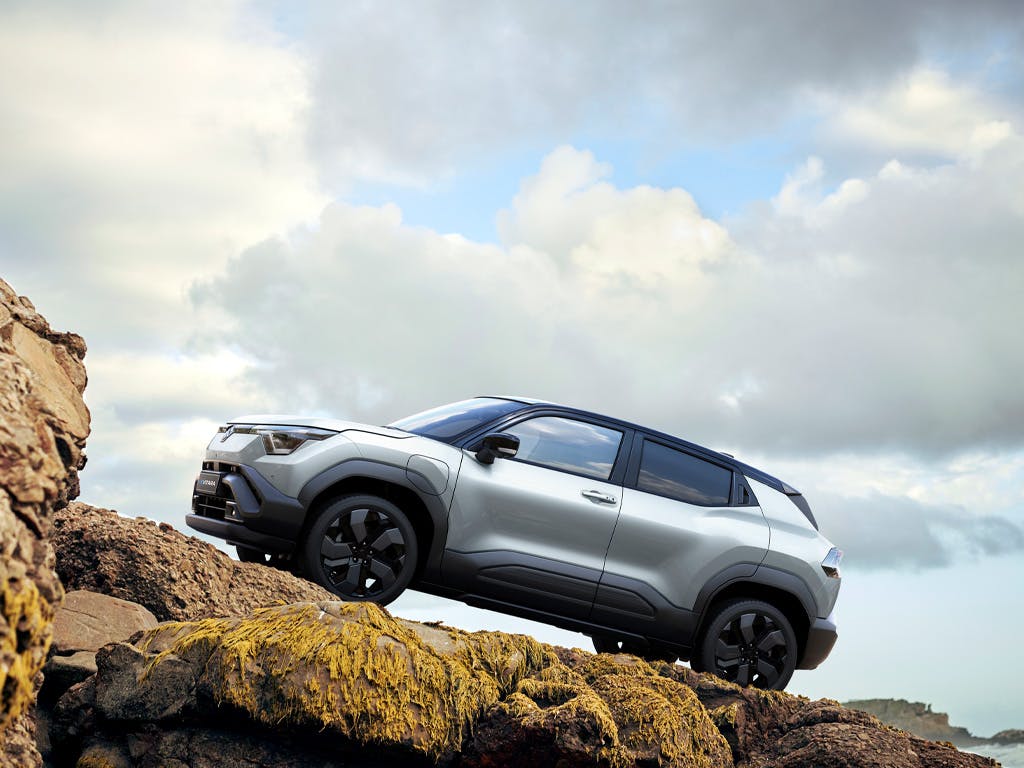 Side profile of the Suzuki e Vitara compact SUV in silver, positioned on rugged coastal rocks showcasing its bold styling, black alloy wheels and raised ground clearance.