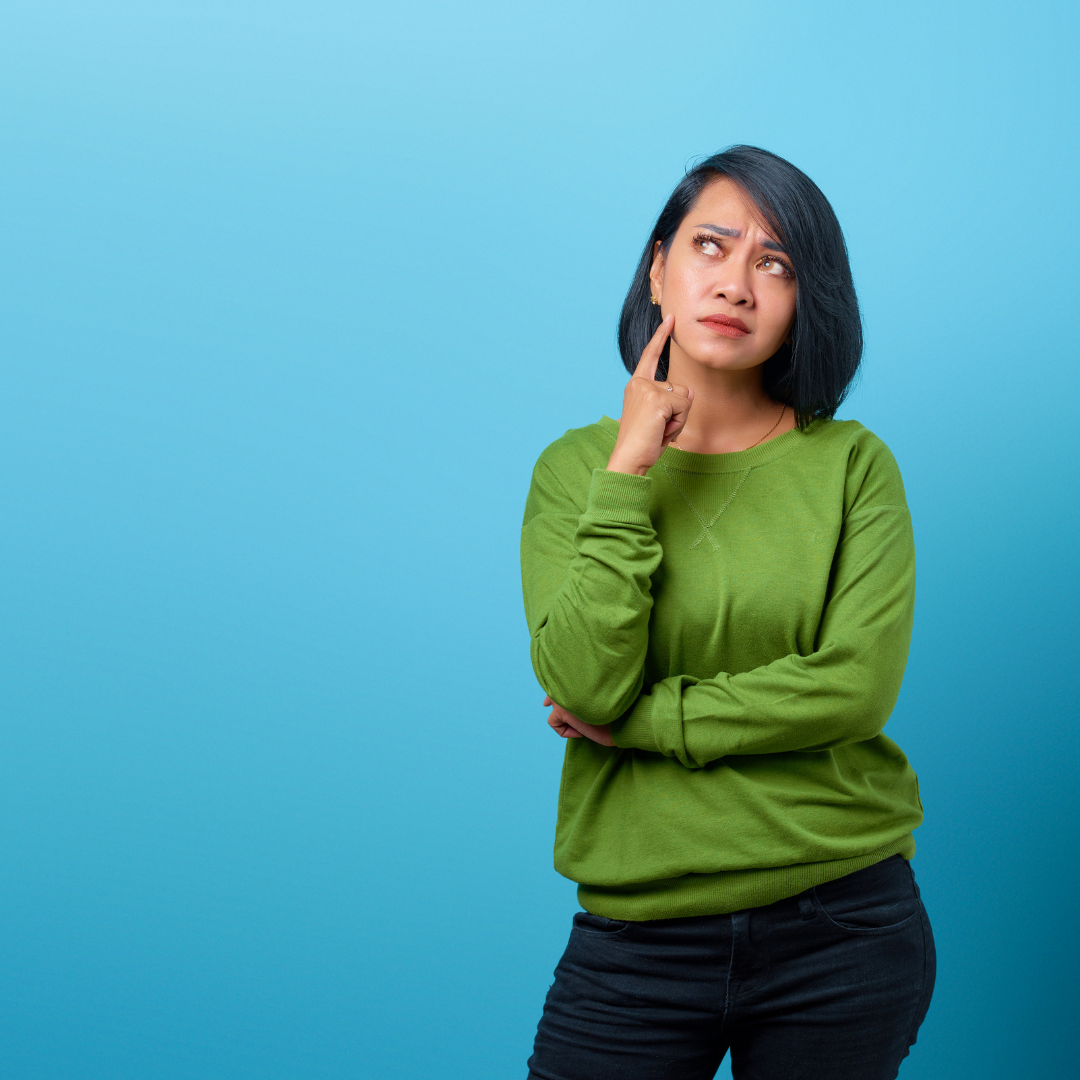 Woman wearing green long sleeve tshirt with black short hair against a blue wall is thinking