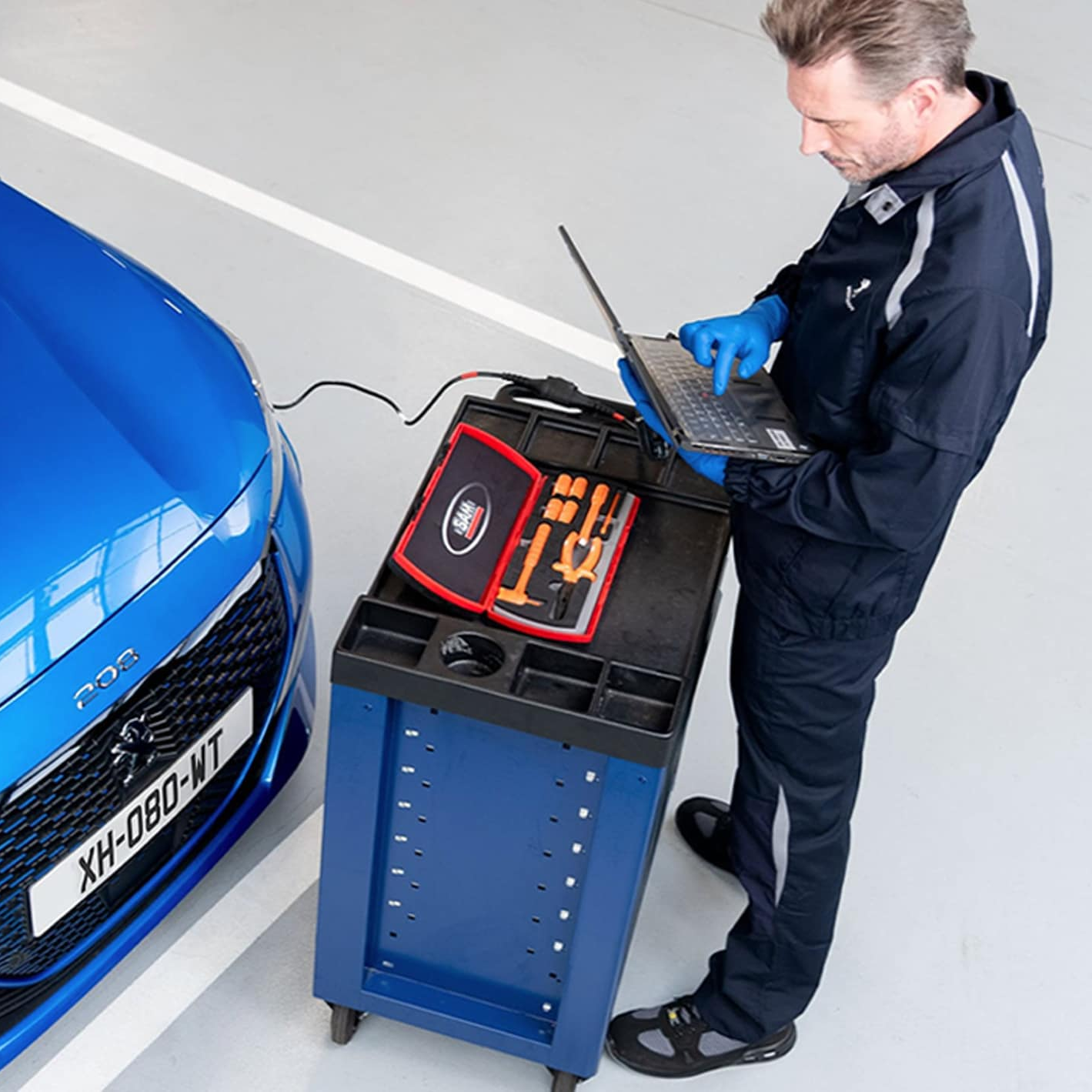 Automotive technician using a laptop beside a blue Peugeot vehicle with a tool trolley containing diagnostic equipment.