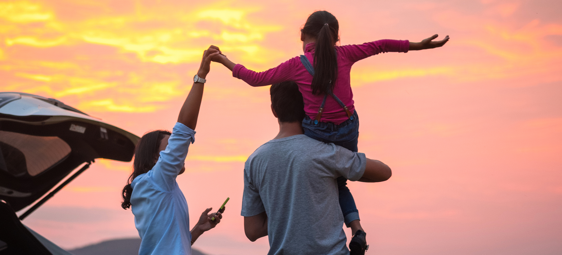 Family with child on dad shoulders next to open car boot