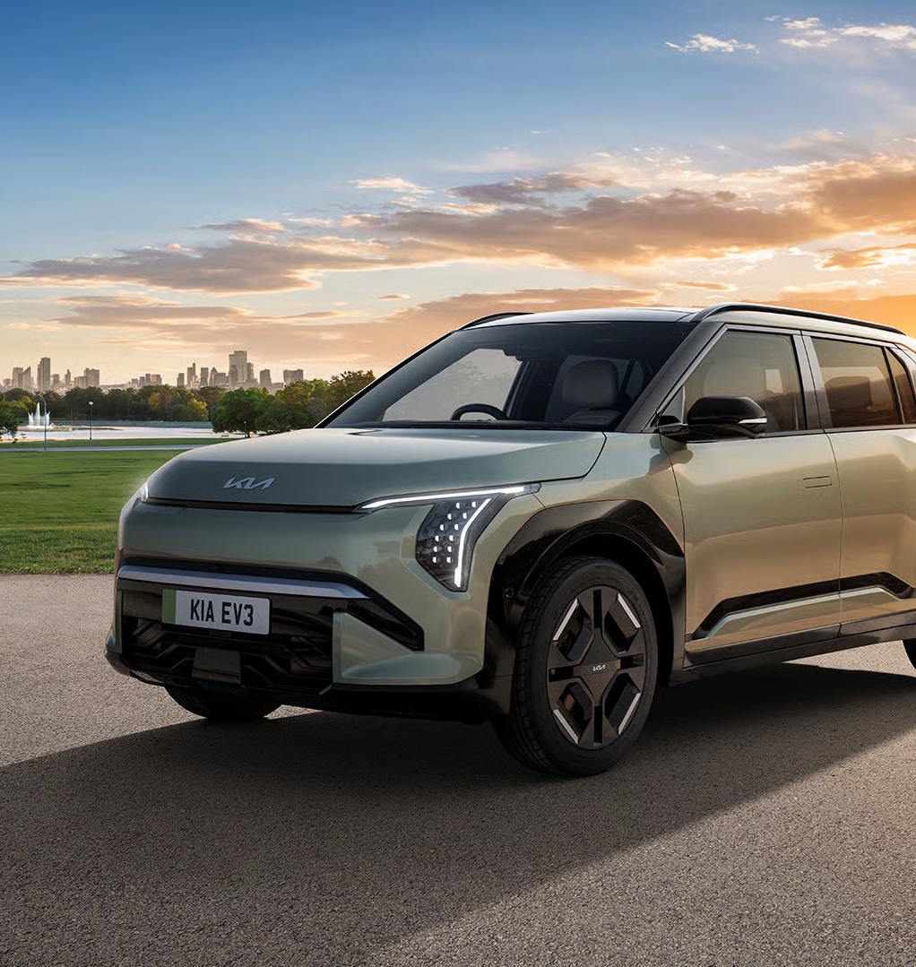 Kia EV3 parked near a park with a city skyline at sunset.