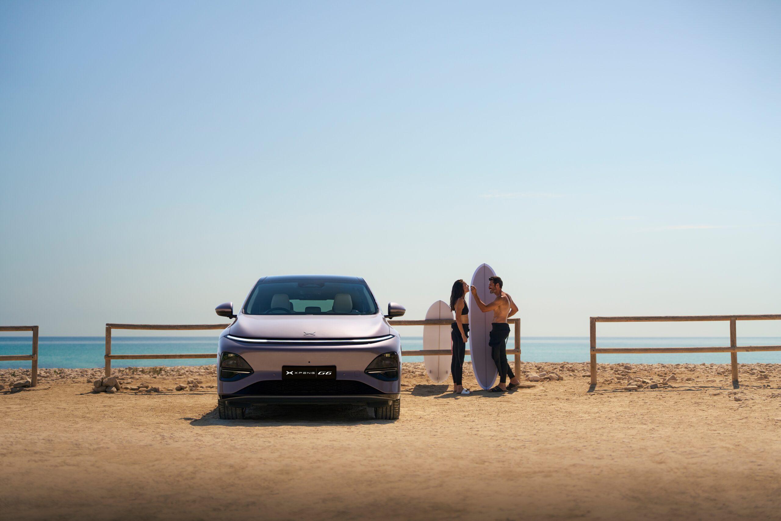 XPeng G6 electric SUV parked on a sandy beach beside two surfers holding surfboards by the sea.