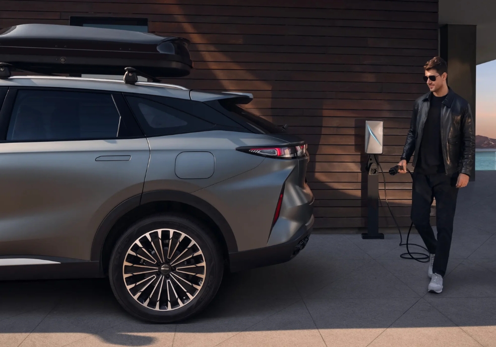 Rear side view of the OMODA 9 parked beside a modern home while a man plugs in the charging cable.