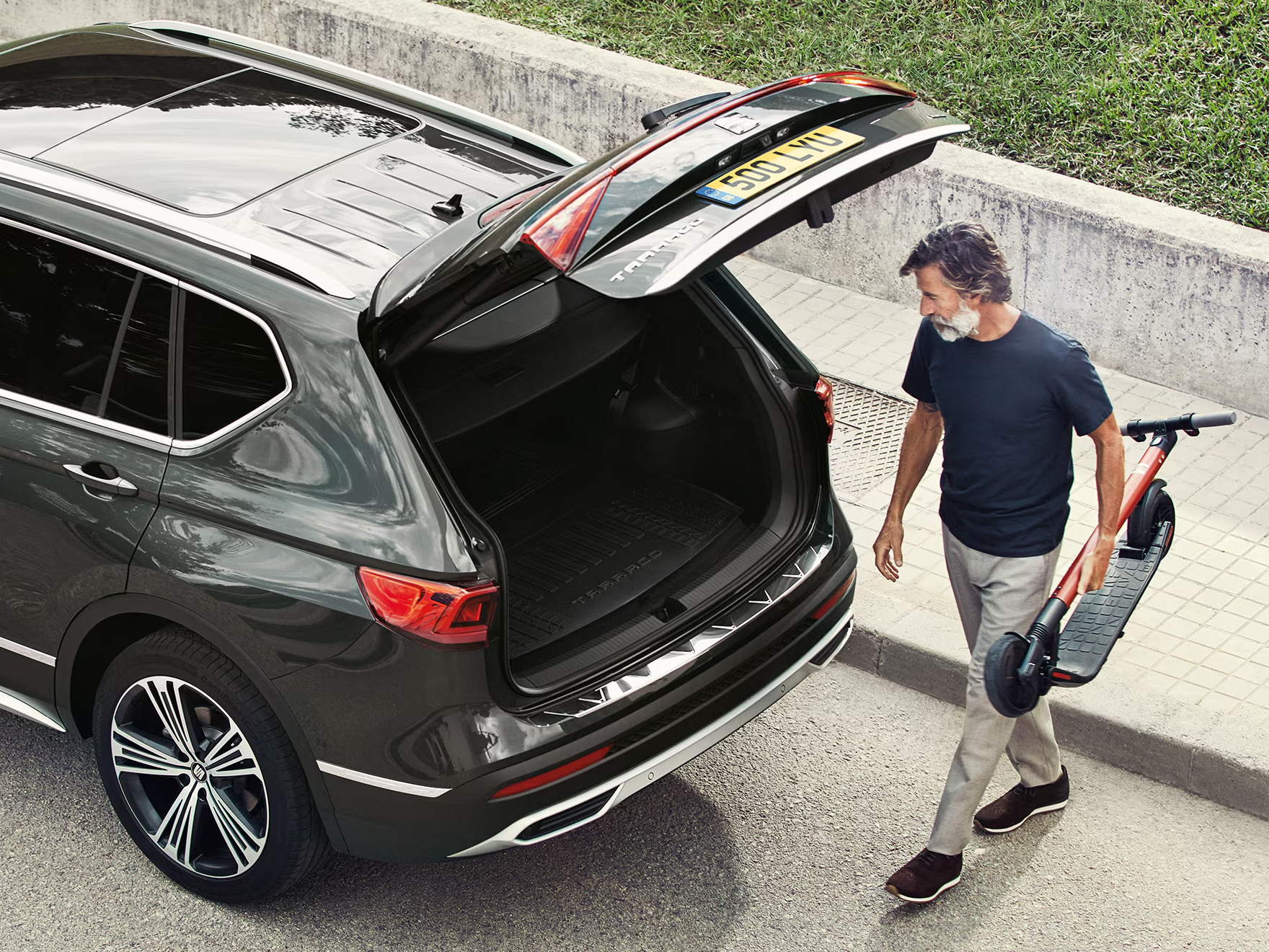 Man walking towards a SEAT Tarraco with the tailgate open, carrying a folded electric scooter ready to be loaded into the boot.