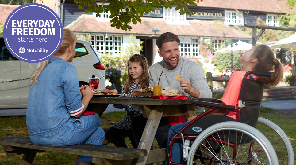 Family laughing whilst eating at a picnic bench. One of the daughters is in a wheelchair. Their motability car is in the background.