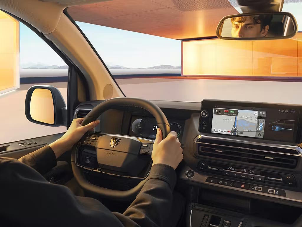 Interior driver's view of the Peugeot E-Traveller, featuring the modern digital instrument cluster, central touchscreen with navigation, and distinctive two-spoke steering wheel with Peugeot emblem.