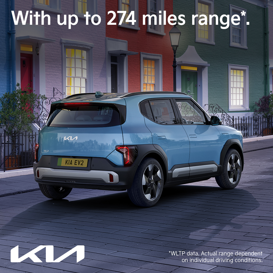 Rear three-quarter view of the Kia EV2 compact electric SUV in blue, parked on a charming urban street, with text highlighting up to 274 miles range.