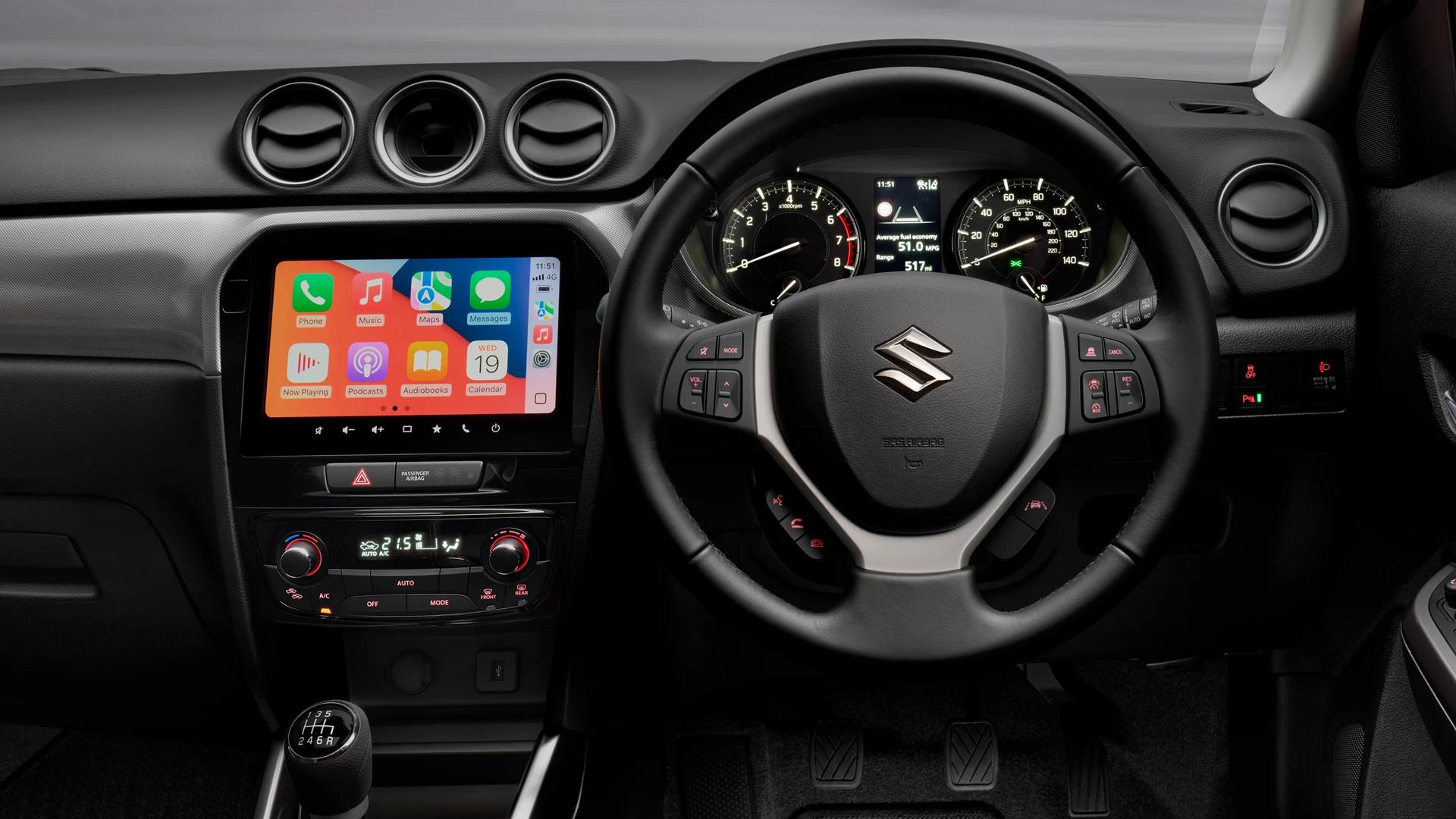 Suzuki car interior showing steering wheel, analogue dials, and central touchscreen with smartphone integration.