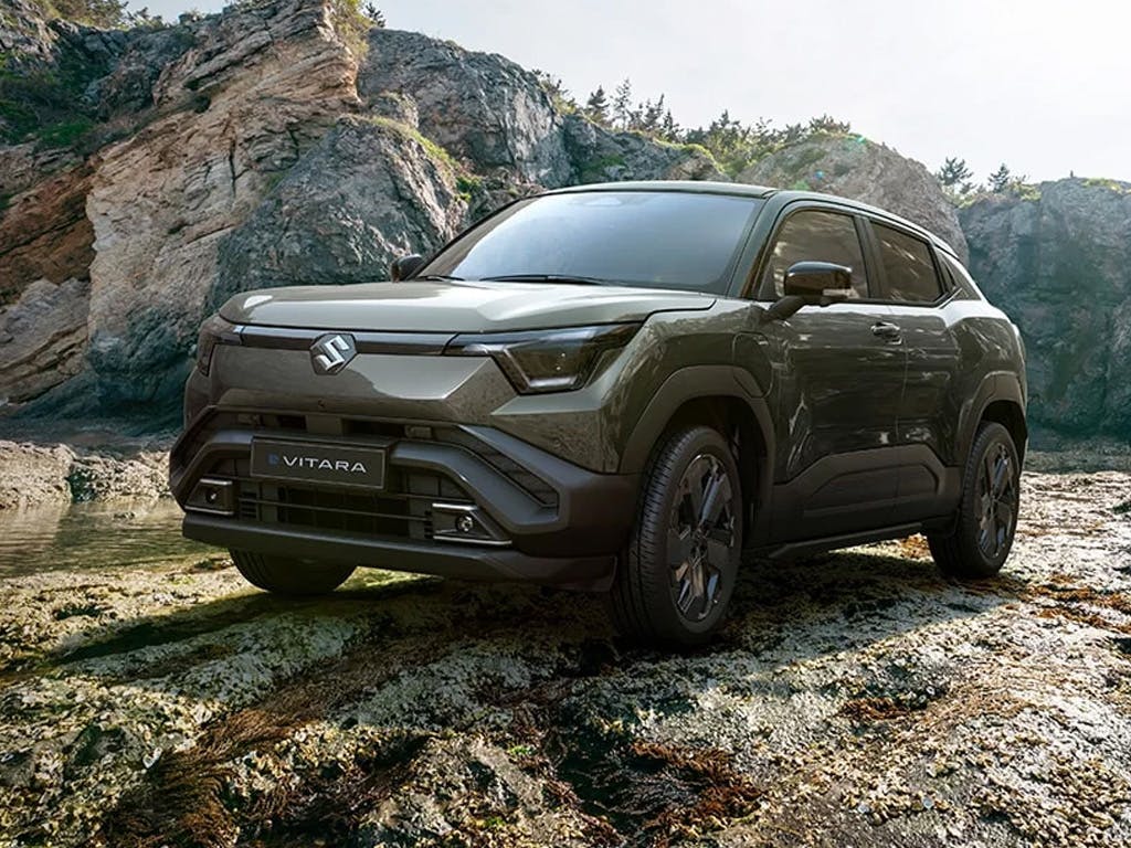 Front three-quarter view of the Suzuki e Vitara compact SUV in olive green, showcasing its bold angular grille, split LED headlights and rugged black body cladding against a rocky coastal backdrop.