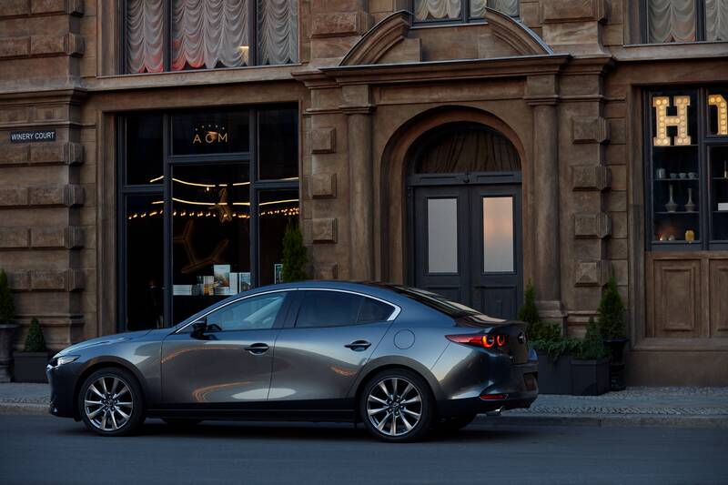 A grey Mazda3 hatchback parked on the side of a road outside a building.