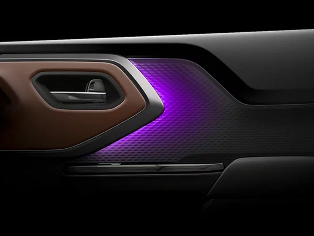 Interior close-up of the Suzuki e Vitara door panel, featuring tan leather trim, chrome door handle surround and striking purple ambient lighting with textured pattern.