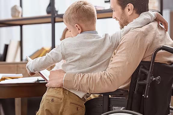 A man in a wheelchair reading a book with a young boy sitting on his lap.
