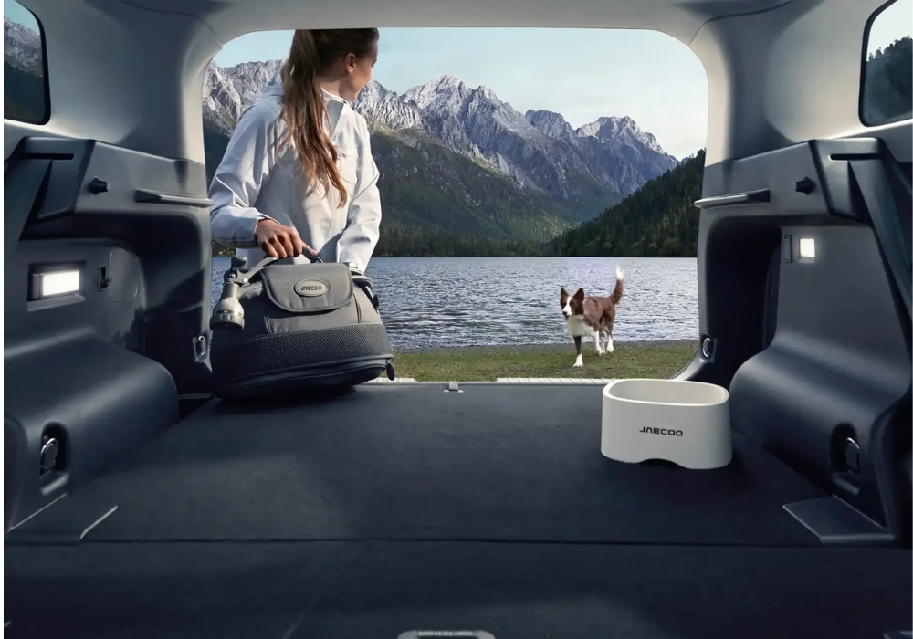 View from inside the open trunk of a Jaecoo 5 showing a bag and pet bowl, with a person and dog by a lakeside in the distance.