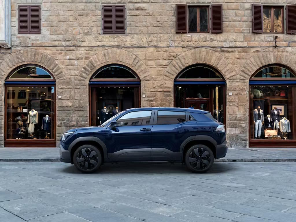 Side profile view of the Suzuki e Vitara compact SUV in deep blue, featuring bold black alloy wheels, rugged body cladding and a sleek crossover silhouette against a historic Italian streetscape.