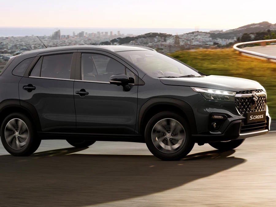 Side profile of the Suzuki S-Cross SUV in dark grey, driving along a coastal road with its bold front grille, LED headlights and sculpted body lines on display.