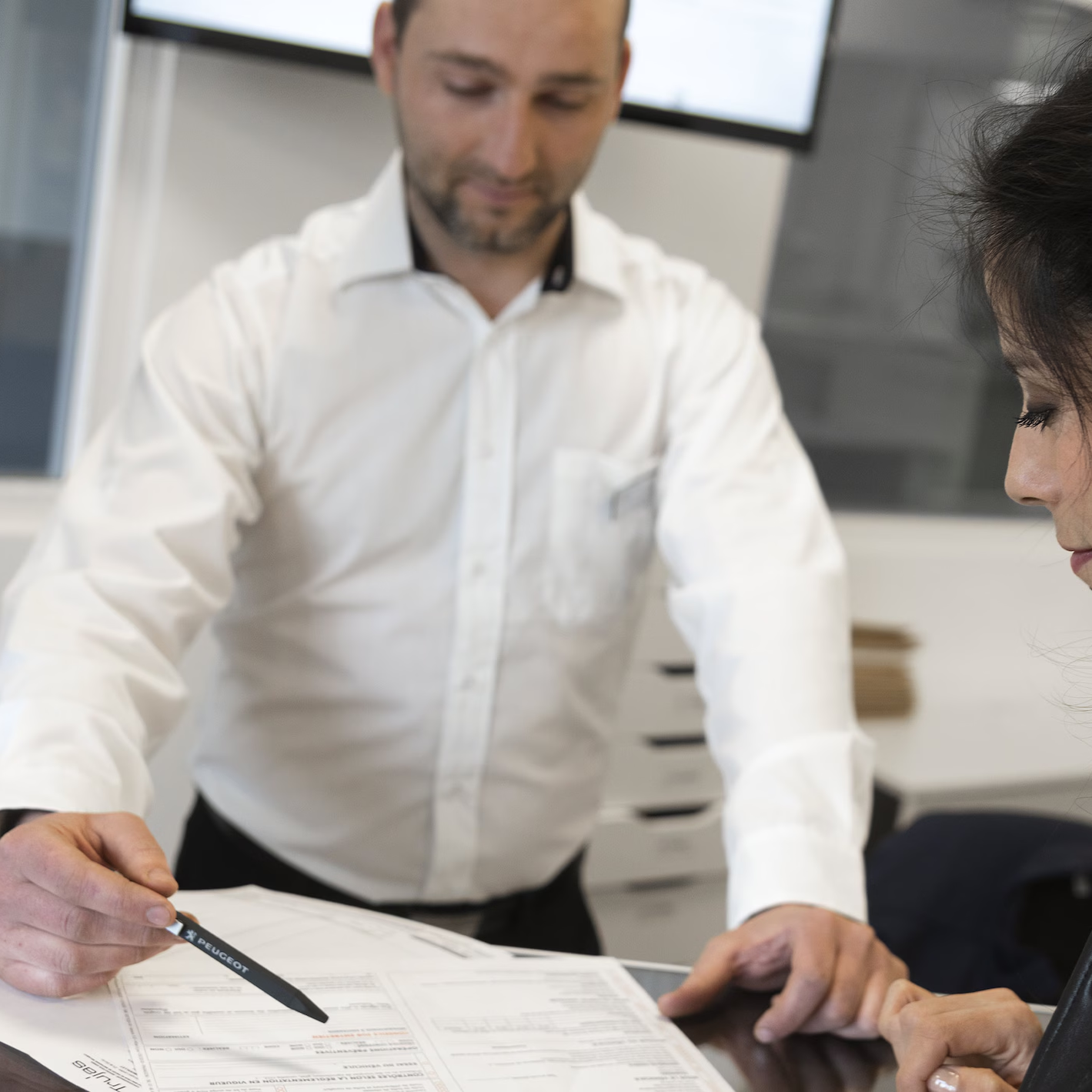 Man in a white shirt reviewing documents and pointing with a pen while assisting a seated customer in an office setting.