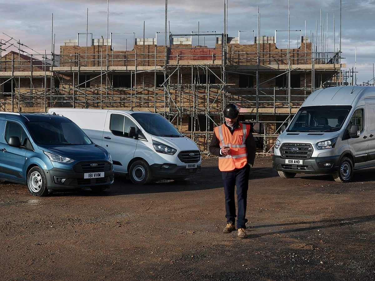A work on a construction site wearing a high vis vest. There are three Ford Transit vans in the background