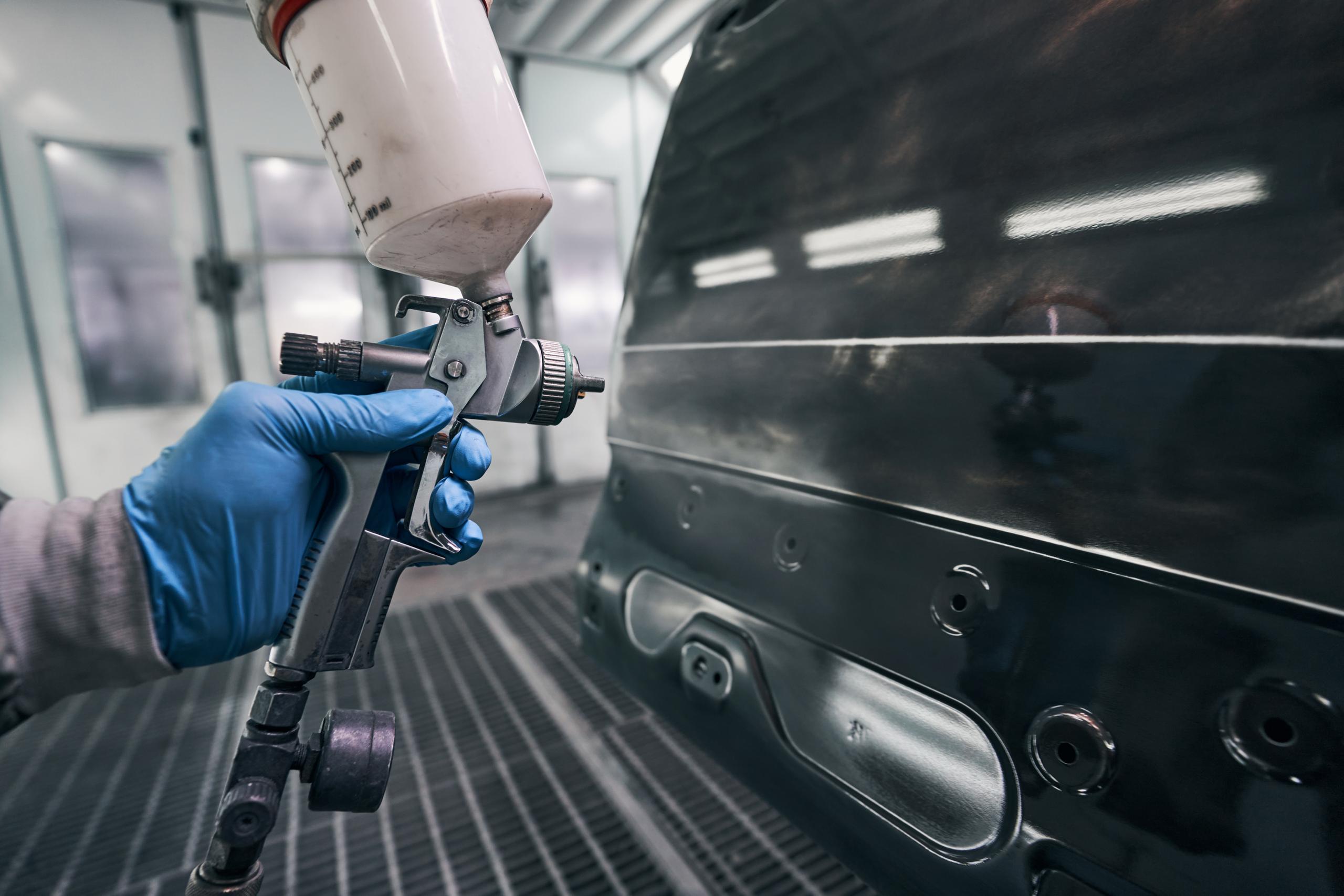 Technician using a spray gun to apply coating to a car panel in a workshop paint booth.