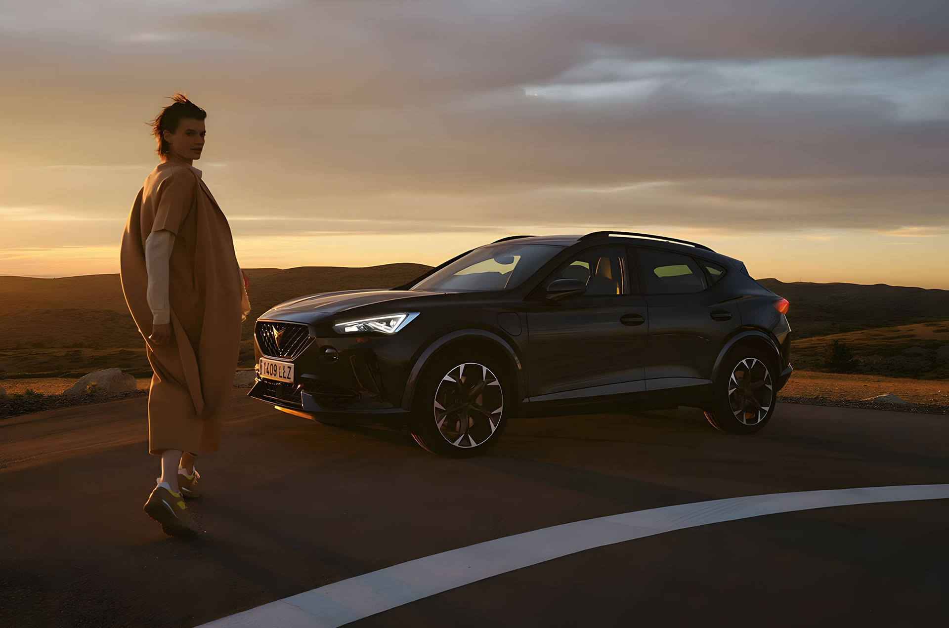 Person walking near a CUPRA Formentor parked on an open road at sunset, with warm light highlighting the SUV’s sleek silhouette against the landscape.
