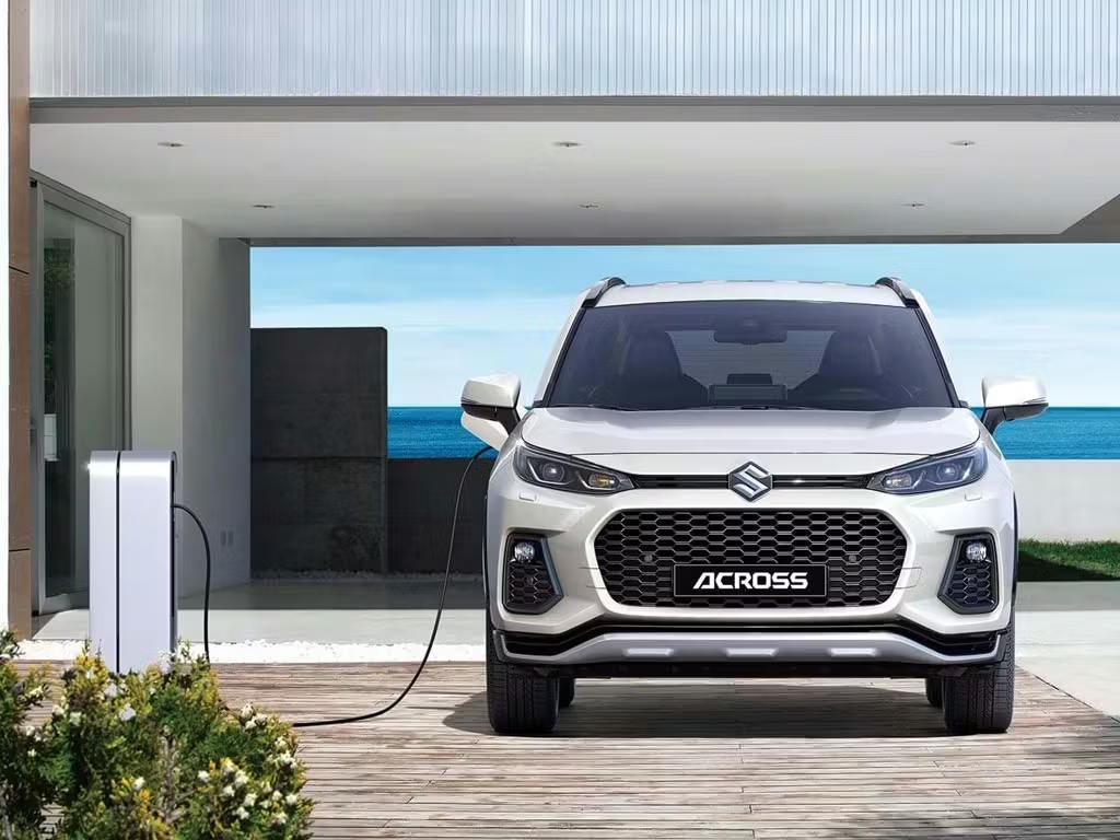 Front view of the white Suzuki Across plug-in hybrid SUV connected to a home charging unit, featuring bold honeycomb grille and sharp LED headlight design.