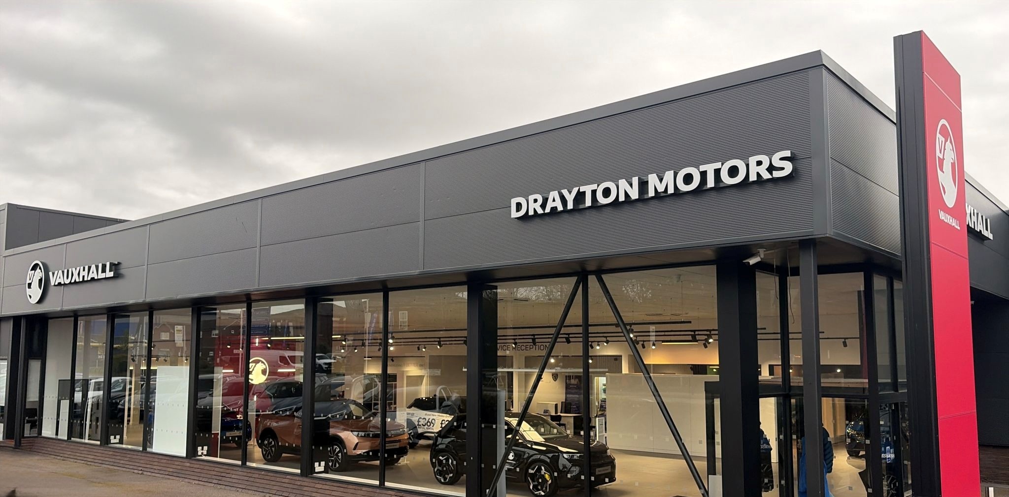 Exterior view of Drayton Motors Vauxhall dealership in Bolton, featuring modern grey cladding, full-height glass showroom windows and prominent Vauxhall branding with vehicles on display inside.