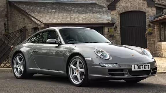 Silver Porsche 911 coupe parked on a driveway in front of stone houses, viewed from the front three‑quarter angle.