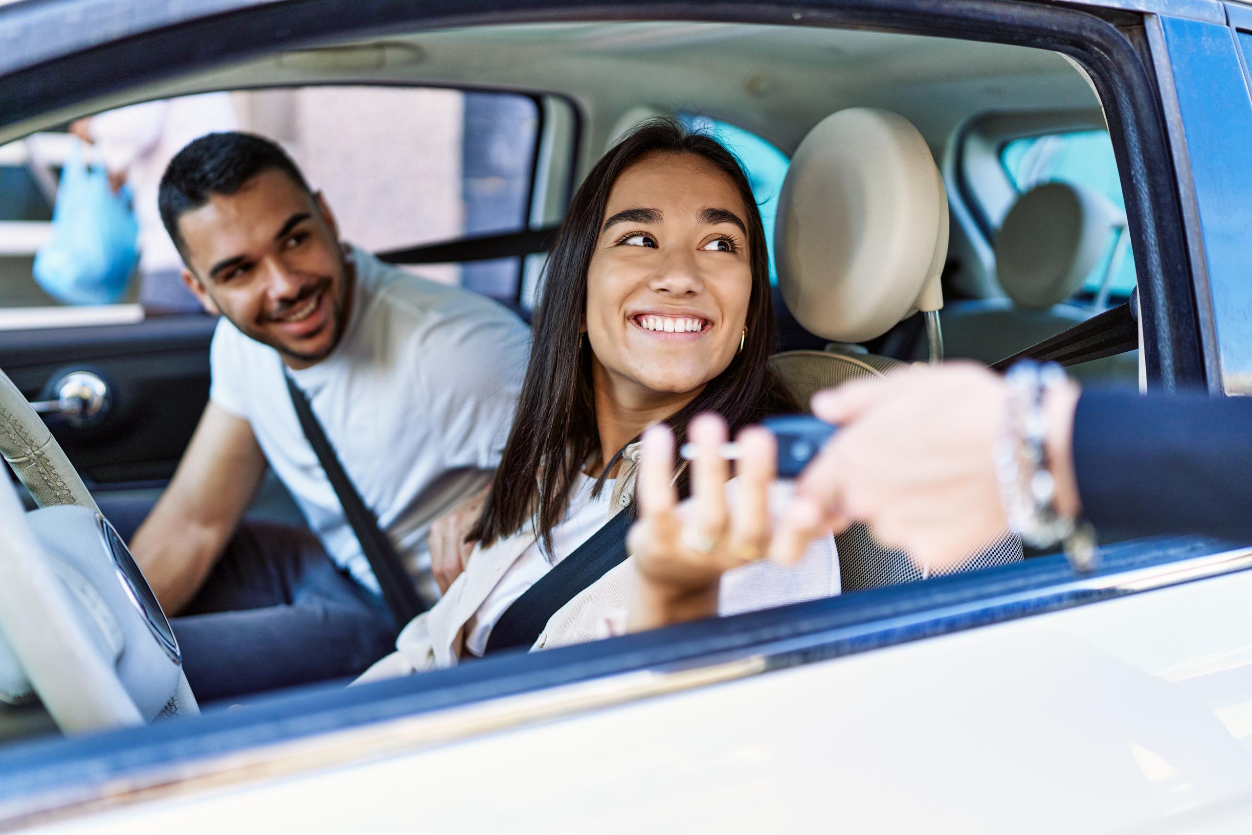 Smiling driver receiving car keys through the window while sitting in a car with a passenger.