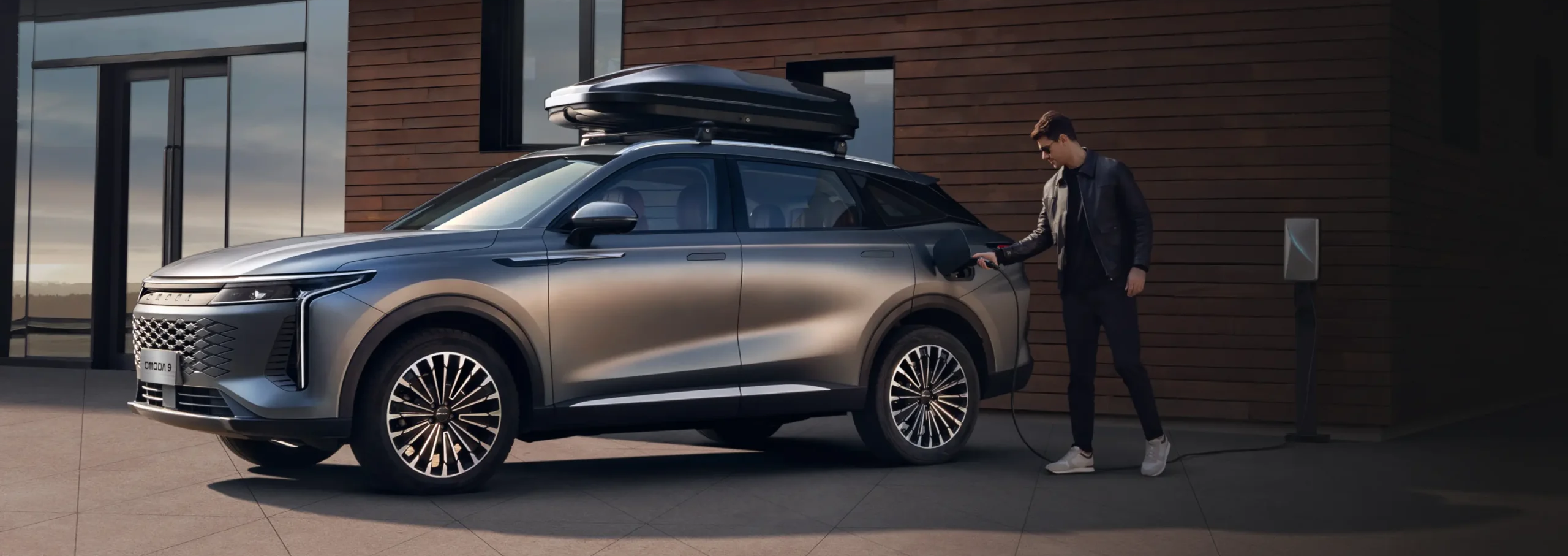 Grey Omoda E5 electric SUV parked outside a modern house with a roof box, as a man plugs in the charging cable.
